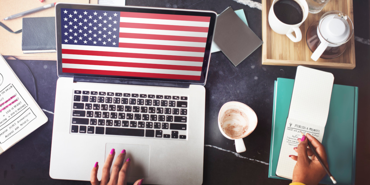 Laptop stojący na biurku, wyświetlający flagę USA. Obok notes, kawa, inne przedmioty.