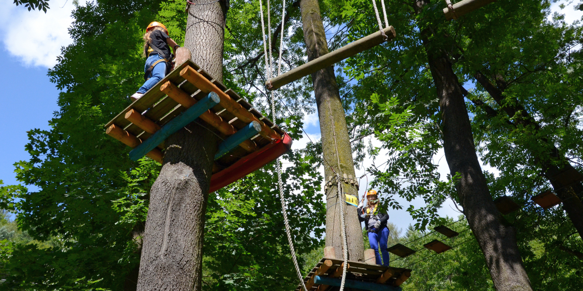 Dwie osoby w kaskach pokonują przeszkody parku linowego, stojąc na drewnianych platformach zawieszonych między drzewami.