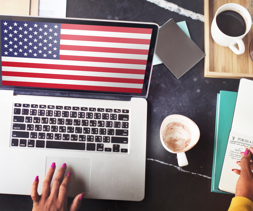 Laptop stojący na biurku, wyświetlający flagę USA. Obok notes, kawa, inne przedmioty. 