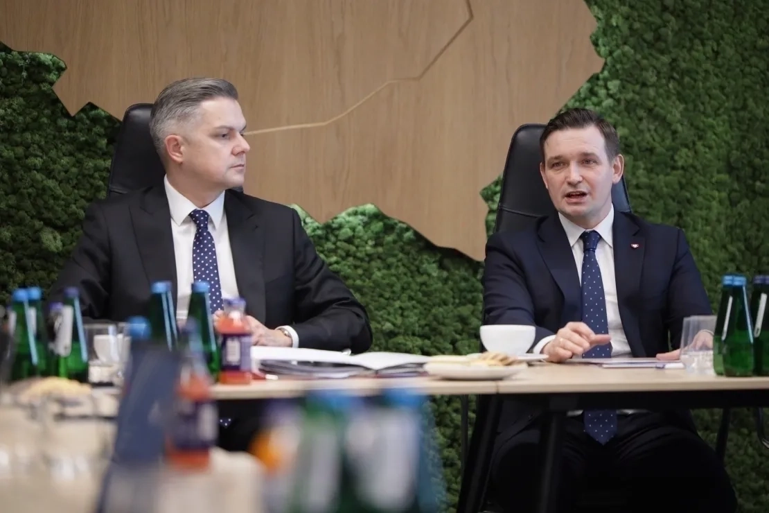 Dwóch mężczyzn siedzących przy stole stojącym przed ścianą z drewnianą mapą